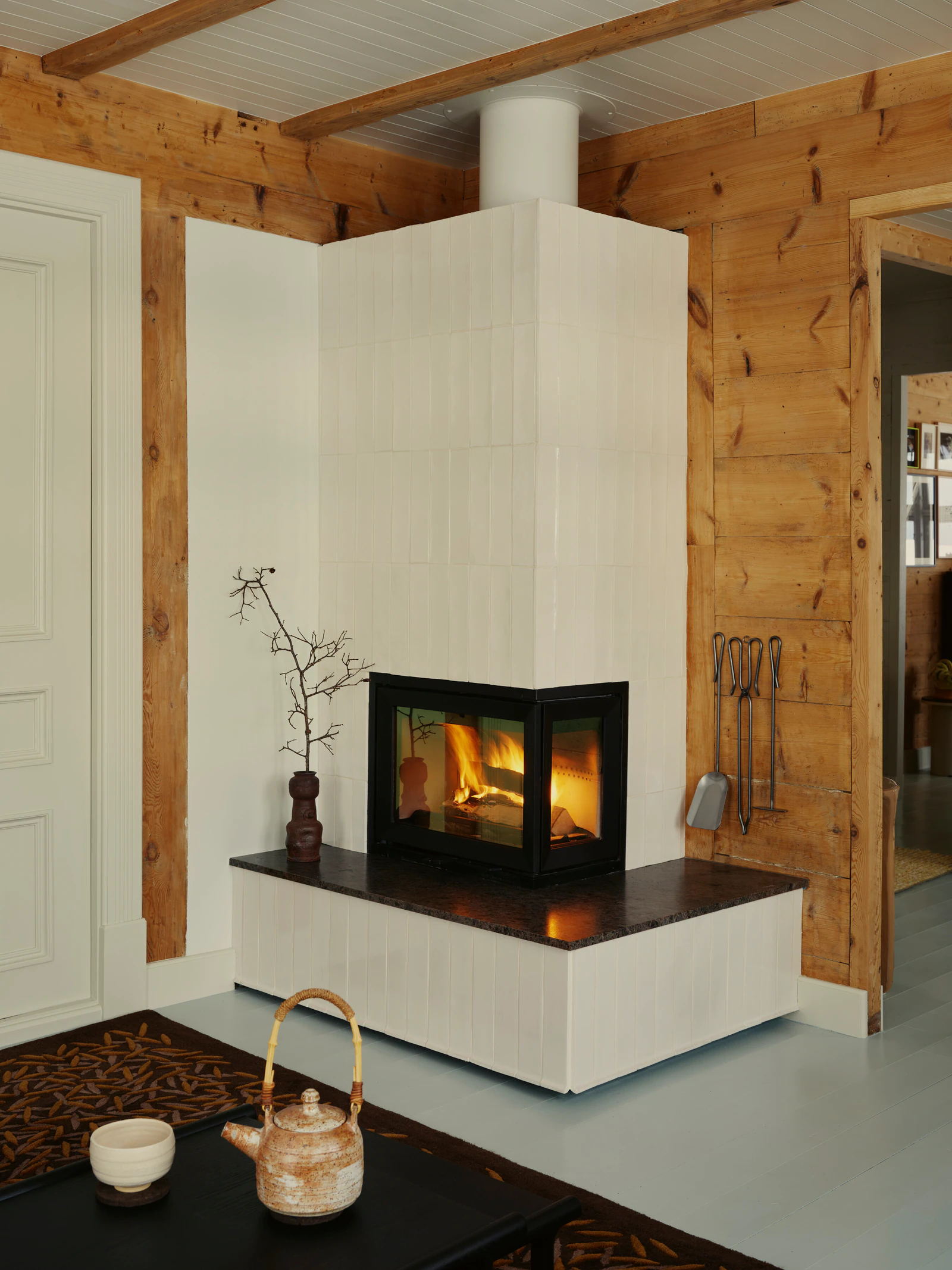 A white-tiled corner fireplace with a fire against a wooden wall, a tea tray with a pot in front.