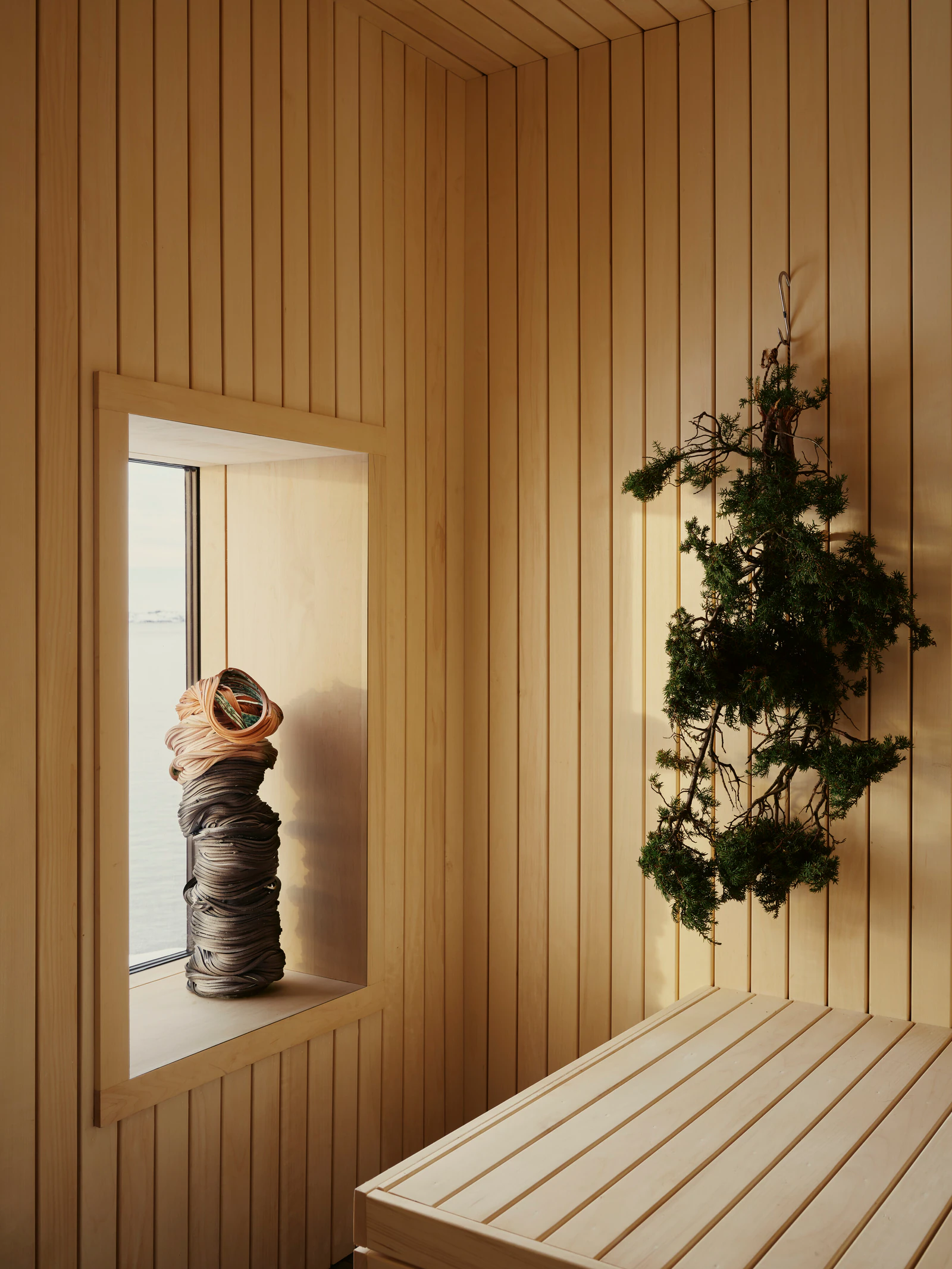 A sauna of pale wood with a window, a bundle of branches hanging on the wall.