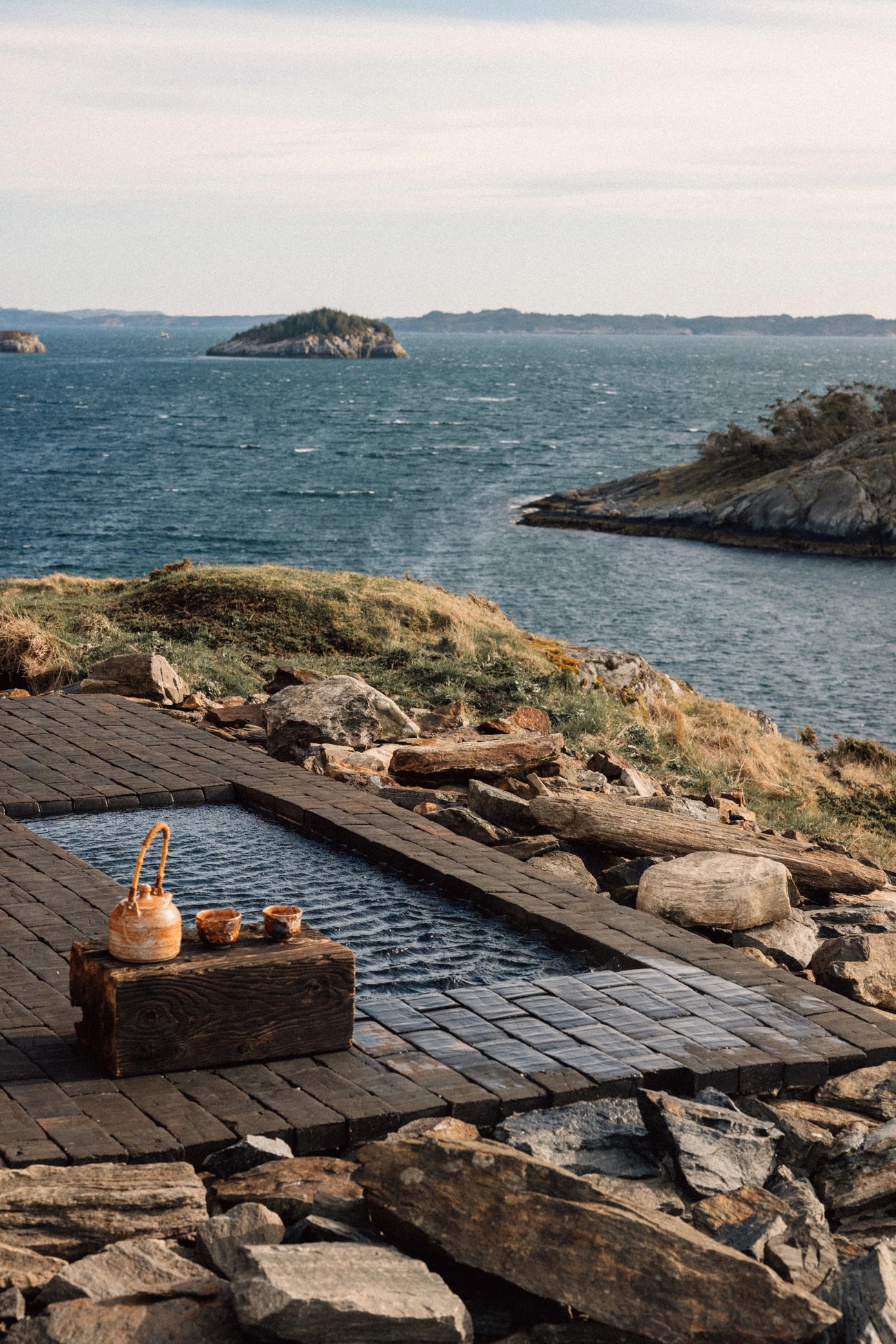 A tub set into the rocks with a timber deck, a kettle on a crate beside it, the sea and small islands beyond.
