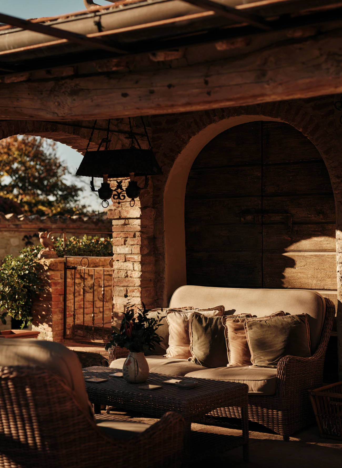 Sitzplatz mit Korbsofa unter der Pergola an einer Ziegelwand.