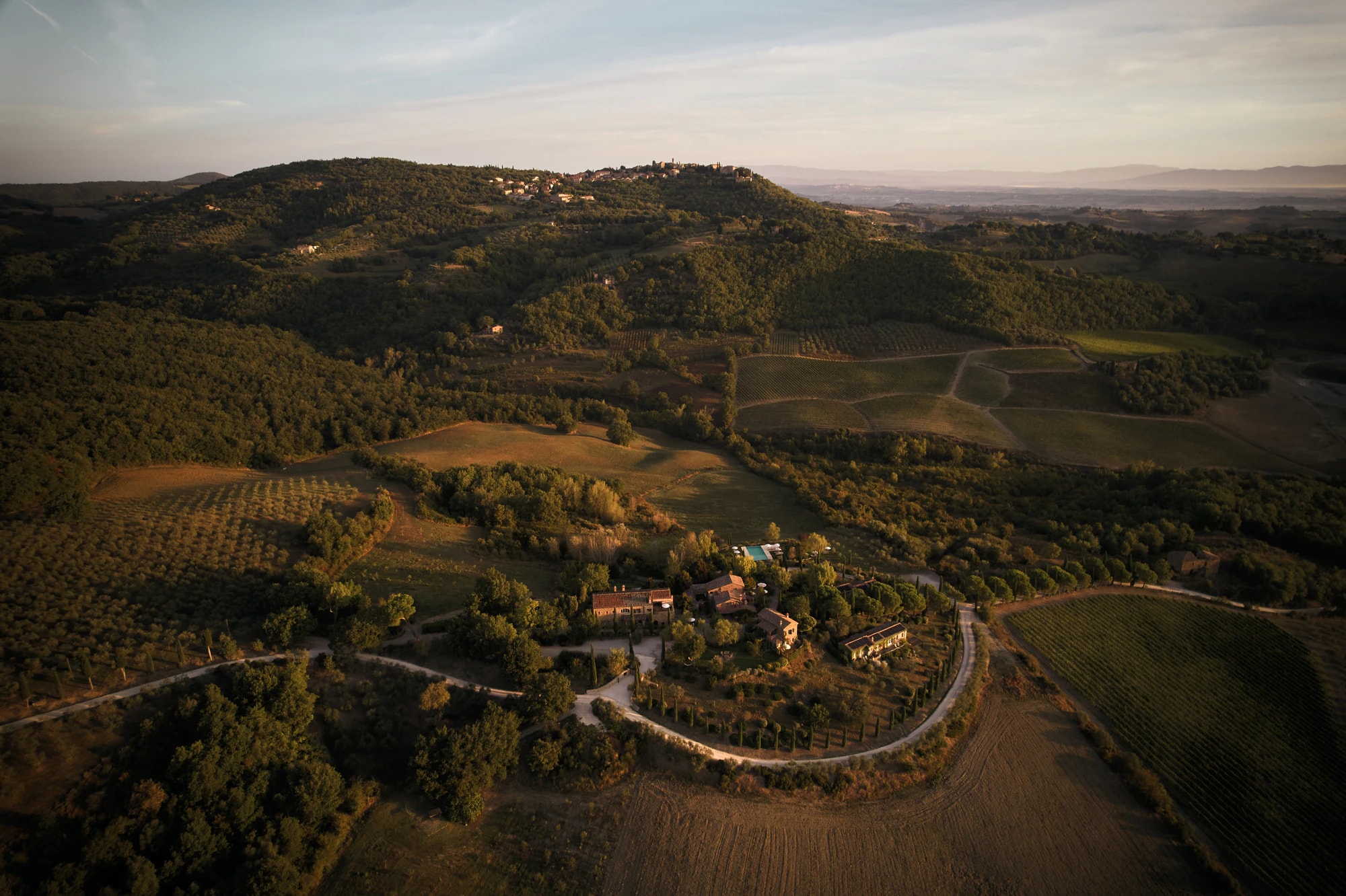 Luftbild des Weilers mit Schotterzufahrt, Olivenhain und Weinberg, dahinter ein Hügelort.