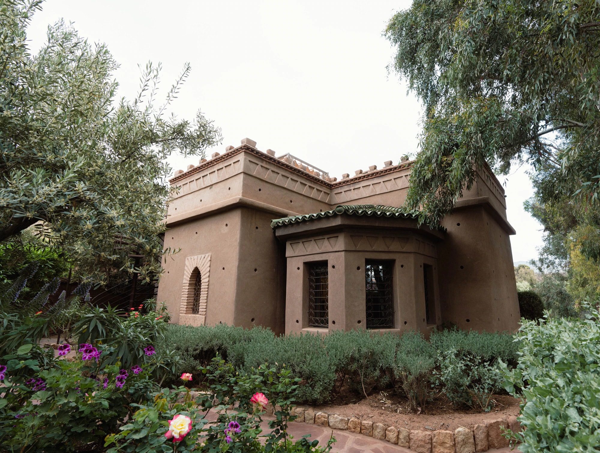 Pavillon aus Lehm inmitten eines Gartens mit Olivenbäumen und blühenden Beeten.