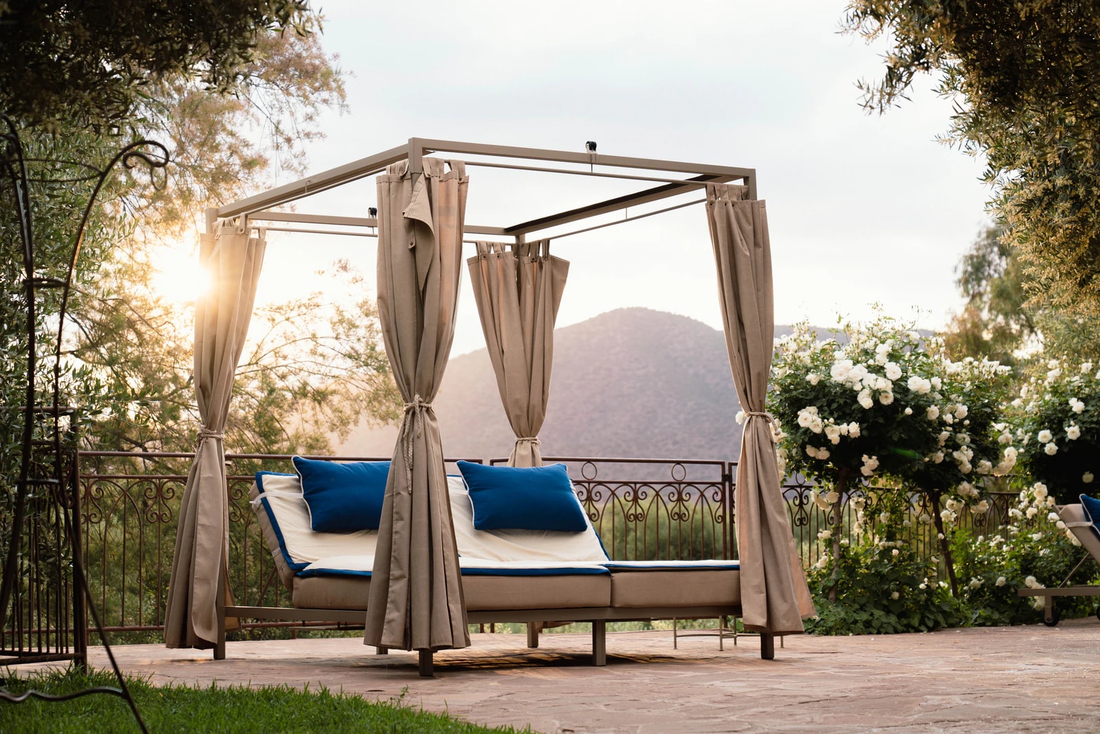 Daybed mit Vorhängen und blauen Kissen am Pool, dahinter Rosen und Berge im Gegenlicht.