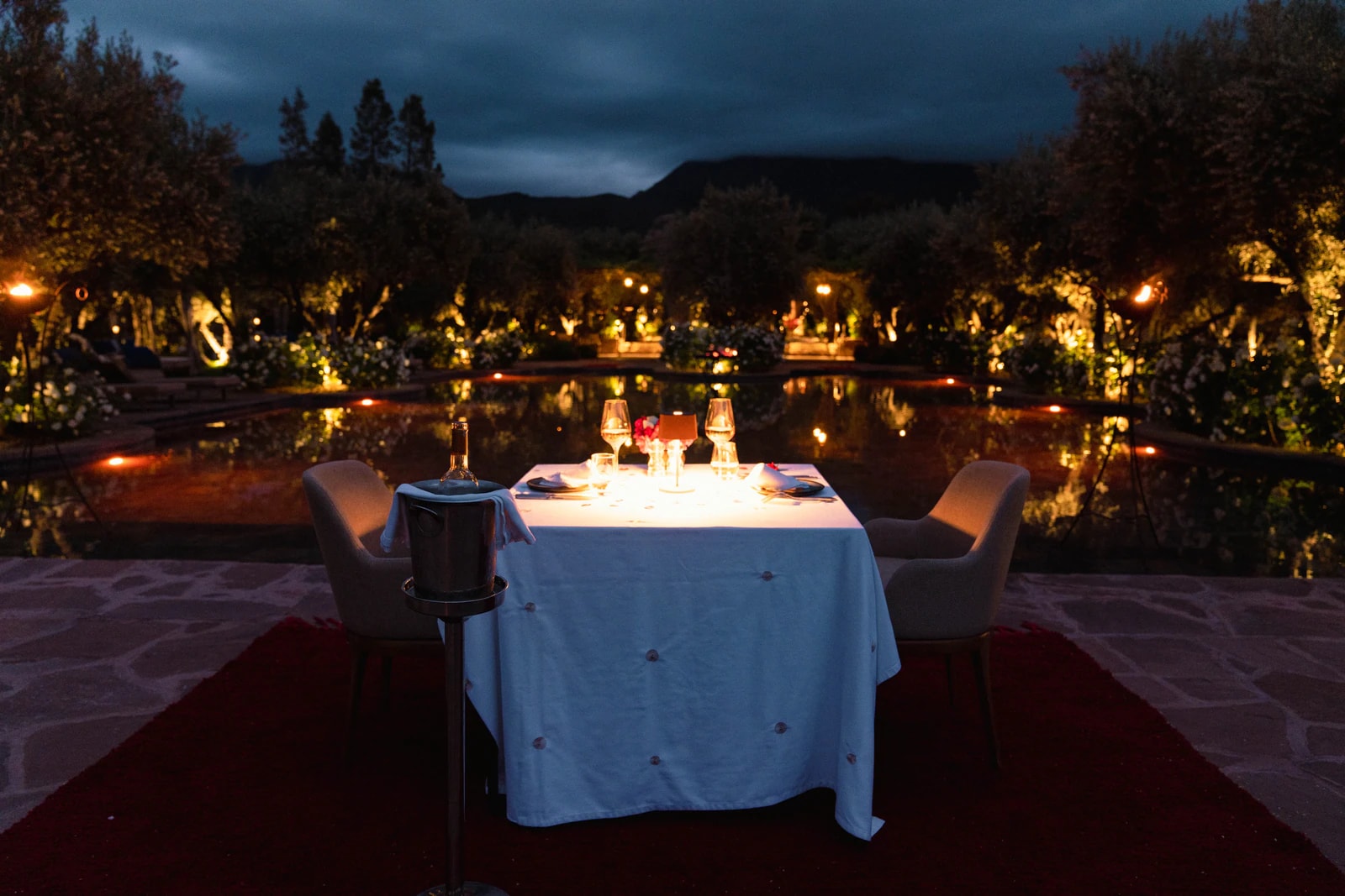 Gedeckter Tisch für zwei am Pool bei Nacht, dahinter beleuchtete Bäume.