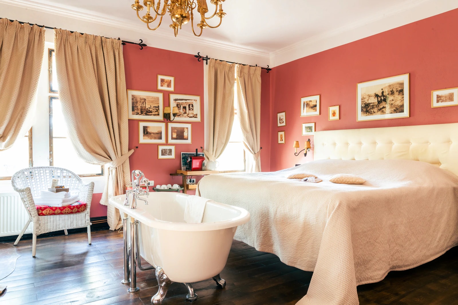 Zimmer mit roten Wänden, gerahmten Bildern, freistehender Badewanne und Doppelbett unter einem Kronleuchter.