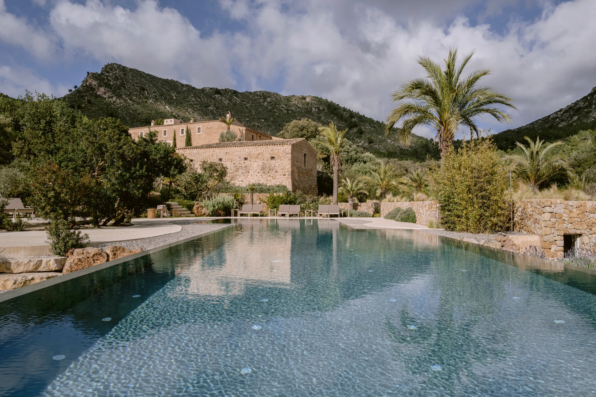 Pool vor den Natursteingebäuden der Finca, dahinter Palmen und ein bewaldeter Berg. Foto: Es Racó d’Artà