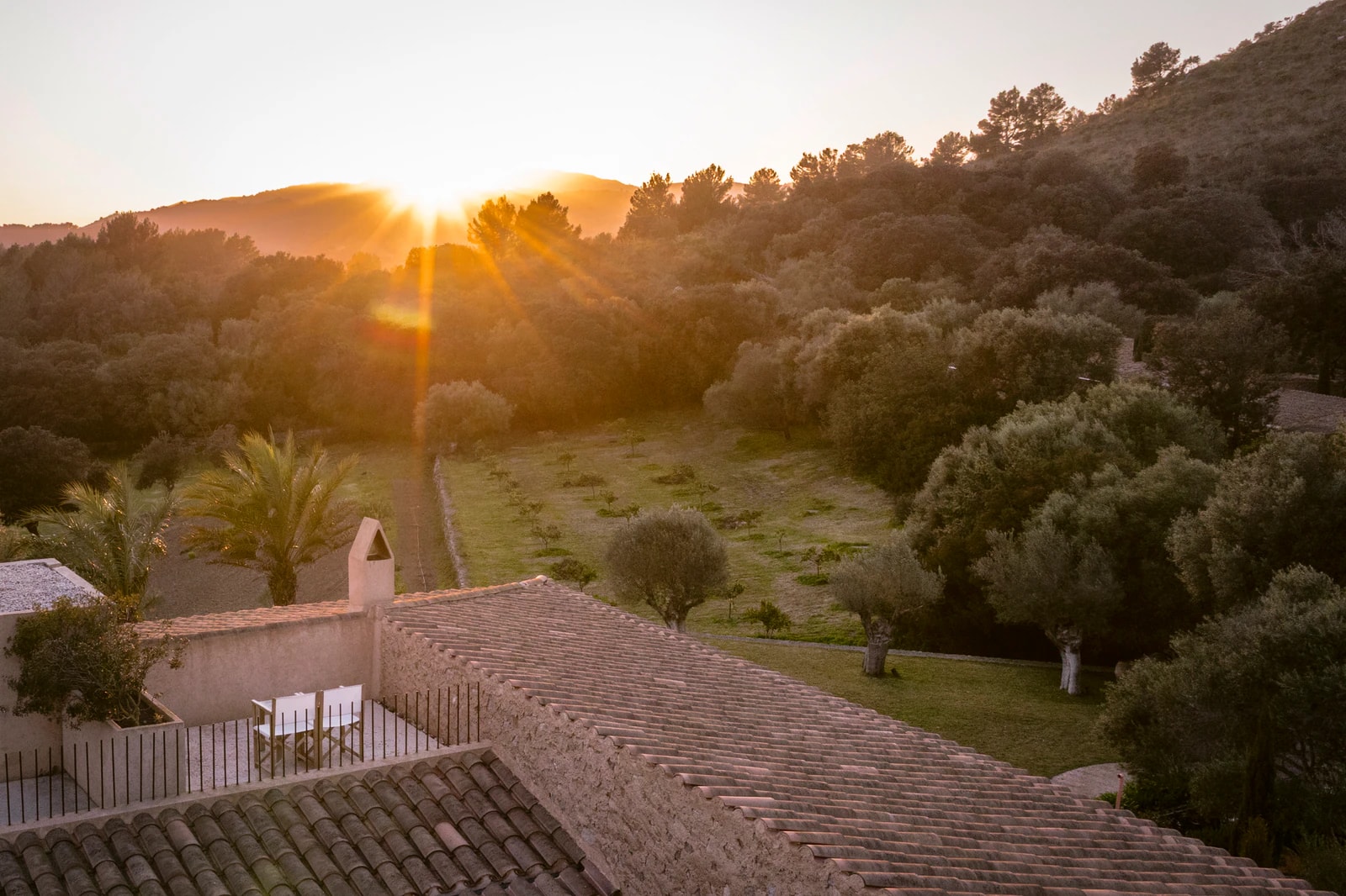 Sonnenuntergang über bewaldeten Hügeln, im Vordergrund Ziegeldächer und eine Terrasse. Foto: Es Racó d’Artà