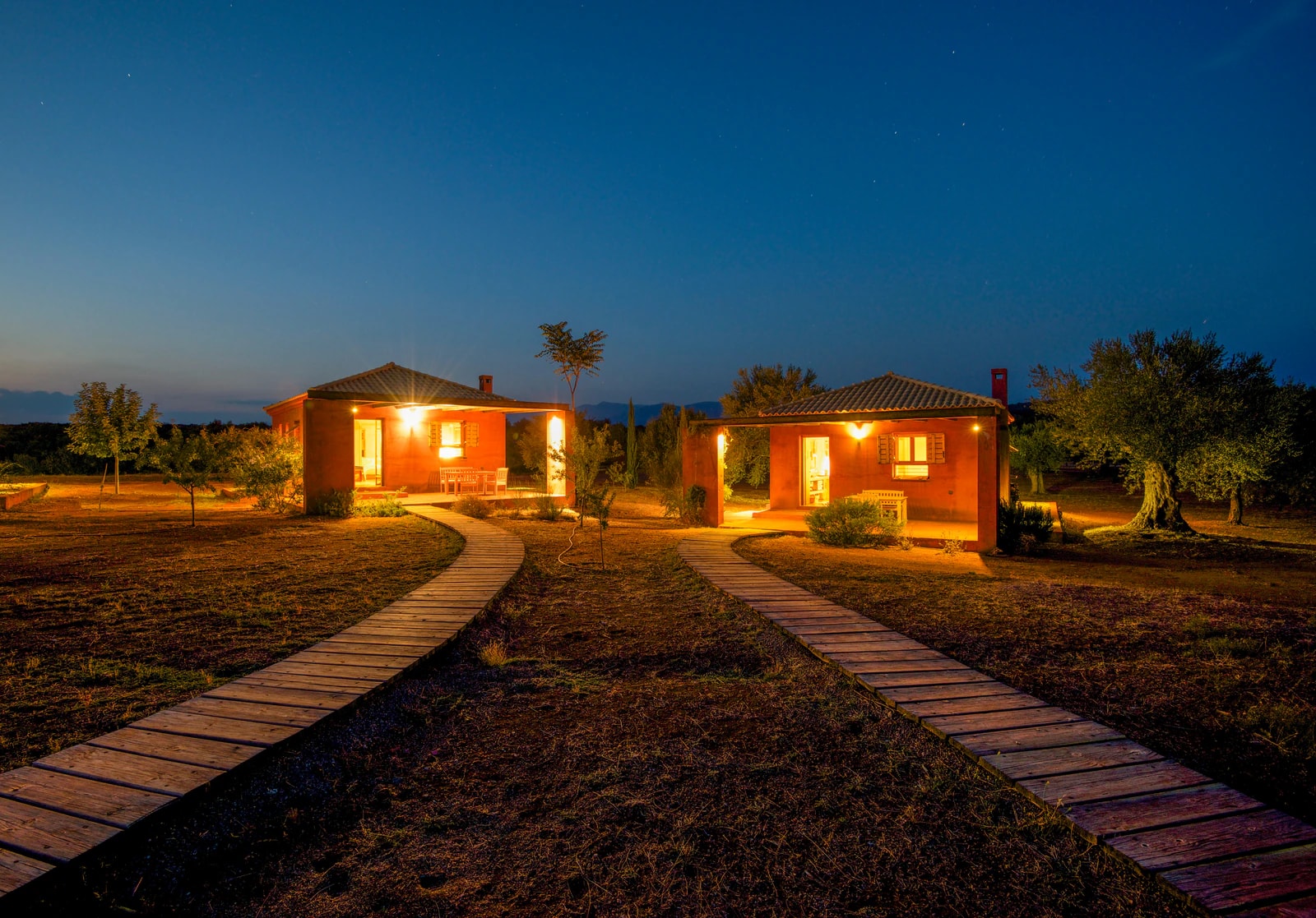 Two lit guest houses with wooden walkways through the meadow at blue hour. Photo: Eumelia